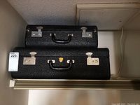 Photo of two black textured vintage suitcases stacked, showing their size difference and metal hardware.