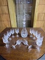 Photo showing 12 crystal wine glasses arranged around 1 crystal decanter with stopper, all placed on a wooden surface with a background of a wood-paneled wall and glass door.