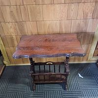 Photo of the antique wooden table with magazine rack against wooden paneled wall, showing scalloped tabletop and vertical slats for magazine storage below.