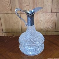 Clear cut crystal liqueur decanter with diamond pattern and silver-tone metal spout and handle against a wooden background.
