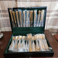 Complete set of gold tone Cambridge flatware including knives, forks, spoons and salad forks in a green velvet-lined black storage case, all wrapped in plastic.