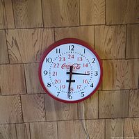 Photo of a round Coca Cola branded wall clock with red frame, white face, dual 12/24 hour markings and electrical cord.