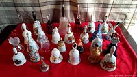 Front view of a red cloth display with 23 assorted collector's bells of various materials and decorations arranged in rows.