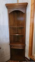 Full view of the wooden corner display cabinet in a room corner, showing its top molding, shelves, and drawer