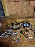 Photo showing assortment of costume jewelry bracelets and bangles on wooden surface, including metal bangles in gold and silver tones, multi-colored beaded bracelets, and chain link bracelets.