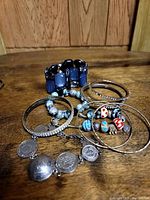 Photo showing multiple bracelets including silver-tone bangles, beaded bracelets with colorful heart and dot patterned beads, and charm bracelets with round medallion charms on a wood surface.