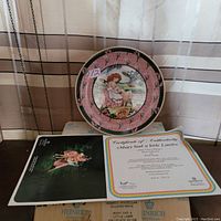 Collector plate facing forward showing illustration of girl with lamb and pink border. Certificate of Authenticity and an informational booklet displayed in front.
