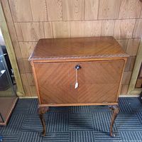 Front view of closed midcentury walnut record cabinet showing carved top edge and decorative legs, with key in keyhole on front.