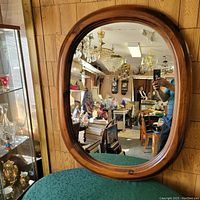 Oval wooden oak frame mirror hanging on paneled wall, showing reflection of a room filled with various antiques and lighting fixtures.