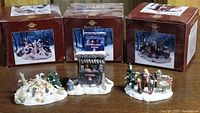Photo of three Norman Rockwell collectible figurines placed in front of their corresponding illustrated boxes showing winter holiday scenes.
