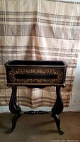 Full front view of antique wooden planter with gold painted floral detailing on black background mounted on curved wooden stand.