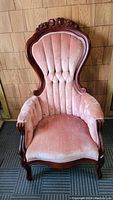 Front view showing the tall, tufted high back and carved wooden frame with floral detail, pink velvet upholstery on the seat and arms.