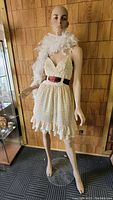 Full body realistic female mannequin standing on a circular clear base wearing a floral dress and white feather boa against a wood-paneled wall.