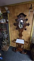 Full front view photo of a walnut Victorian hall tree featuring an oval mirror at the top, a white marble inset console table in the middle section, and multiple hanger and umbrella holders at the bottom. The height is substantial, reaching approximately 89 inches tall. The piece is placed against a wood-paneled wall with other antique items nearby.