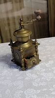 Photo showing vintage brass manual coffee grinder with wooden handle and small drawer for grounds, placed on white lace tablecloth.