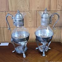 Two vintage glass coffee carafes in silver plated warmer stands on wooden surface.