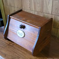 Wooden bread box with a trapezoidal shape, hinged front lid, wooden knob handle, and a decorative 'Bread' label on the front.