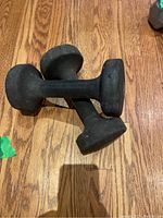 Two black dumbbells placed crossed on a wooden floor, showing the shape and grip area.