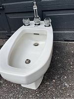 Side angle view of white American Standard bidet showing oval bowl, chrome and acrylic faucet handles, and integrated pedestal base.