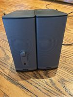 Front view of two black Bose mini speakers side by side showing gray mesh speaker grilles and control knob on front of one unit.
