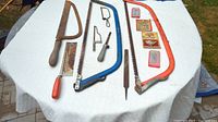 Photo of three hacksaws laid out flat with wooden, blue, and orange handles, additional hand saw, file, and small tools on white tablecloth outdoors.