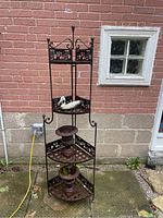 Full view of tall black cast iron corner shelf with three triangular tiers and decorative elephant panels, placed against an outdoor brick wall.