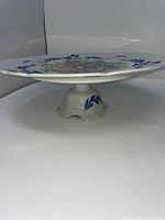 Side angle view of white porcelain cake plate on pedestal, showing blue leaf decoration on foot and overall shape