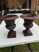 Pair of rusted cast iron urns on a white table outdoors, showing full front view.