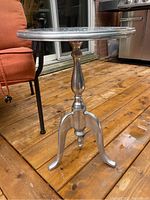 Side view of the galvanized metal pedestal side table on wooden deck floor showing the turned central pillar and three curved legs.
