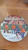 Top down view of hand decorated ceramic plate depicting street hockey game scene, players in orange and blue with buildings and snow background