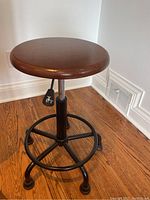 Full view of the retro style stool with round wooden top, metal black base with footrest, and height adjustment lever.