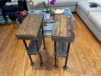 Two industrial side tables shown from side angle on hardwood floor with couch and glass coffee table background.