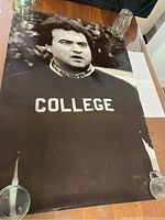 Full collected view of John Belushi black and white poster showing face and COLLEGE sweatshirt, on table with glasses holding corners down.