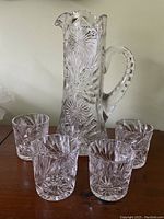 Tall cut glass pitcher with detailed ornate cut patterns and a large handle, alongside four matching cut glass tumblers with geometric starburst and fan etching.