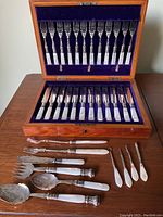 Wooden presentation box open showing 12 dinner forks in top tray, 12 dinner knives in bottom tray, and additional serving pieces displayed in front