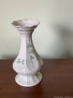Front view of the white Belleek vase with green shamrocks on a wooden surface against a plain wall.