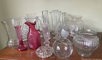 Collection of cranberry glass and cut glass vases displayed on wooden surface, showing variety of sizes, colors, and styles.