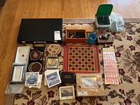 Overview photo showing the full game lot including chess set, ceramic dishes, coasters, marbles, cards, sticker sheets, and game boards laid out on a patterned carpet.