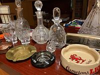 Photo showing multiple crystal decanters with stoppers, two crystal glasses, brass and metal ashtrays, and a ceramic ashtray with Camerons L.I.O. lion logo.