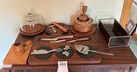 Full display of teak kitchen and bar accessories on a wooden surface including cutting boards, knives, and serving plates