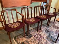 Four walnut dining chairs in a row, leftmost with arms, all upholstered in purple floral needlepoint.