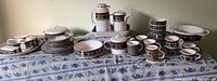 Full display of vintage Mikasa Intaglio pattern dish set including plates, bowls, cups, teapots, serving dishes and tablecloth on blue floral fabric