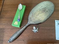 Full view of the three items: sterling silver hand mirror, souvenir spoon in green box, and maple leaf brooch on wooden surface.