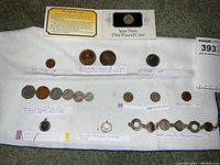 All coins and jewelry laid out on grey carpet with notes indicating coin types and dates such as British Farthing pendant and Victorian coins.