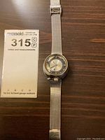 Full view of the vintage MCM space age wristwatch on a wooden surface with lot number card.