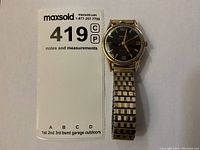 Front view showing black dial, gold-tone case, expansion band and lot tag