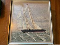 Full view of the needlepoint artwork showing the Bluenose schooner with full sails on the ocean within a thin metal frame and glass covering.