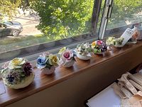 Seven porcelain flower arrangements displayed on a window ledge, showing a variety of floral designs and arrangements from afar to give an overview of the lot.