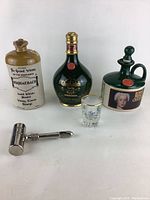 Three ceramic vintage whisky bottles and a clear shot glass arranged on white background with metal tap handle opener.
