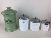 Four bathroom canisters in a row. Left is a tall green ceramic canister with Paris text and three white ceramic canisters labeled No 3, No 2, No 1 with chrome lids.